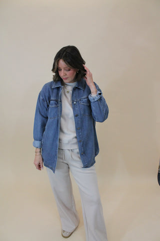Person wearing a blue denim jacket over a white shirt and pants on a beige background